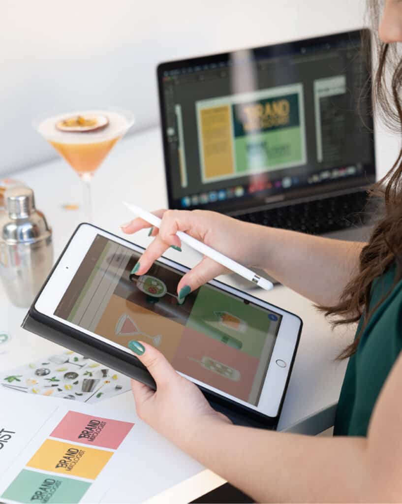 graphic designer woman close up drawing on an ipad their brand icons, on a white desk with a passionfruit cocktail and a computer in the background