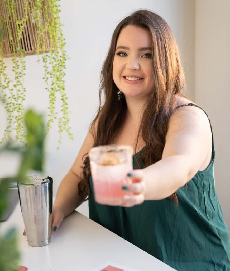 graphic designer pouring a pink margarita cocktail on a desk with a computer in the background and some dried fruits